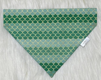 May include: Green and gold dog bandana with a mermaid scale pattern. The bandana has a small white tag with the text "verly's creations" on it.