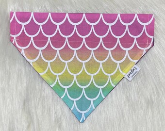 May include: A rainbow ombre dog bandana with a mermaid scale pattern. The bandana has a white border and a small tag that says "Simba's Creations".