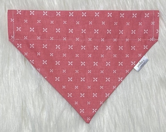May include: A pink dog bandana with a white geometric pattern. The bandana has a white tag with the text "sunbe's creations" on it.