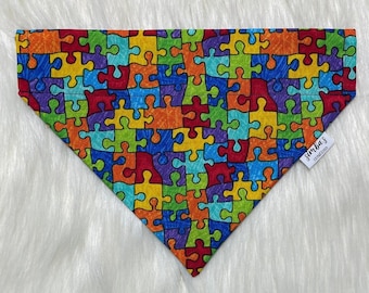 May include: Triangular dog bandana with a colorful puzzle piece pattern. The bandana features a vibrant array of colors including red, orange, yellow, green, blue, and purple. A small white tag with black text is attached to the bandana.