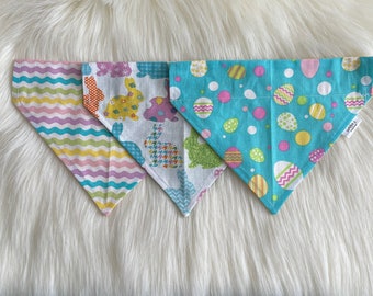 May include: Three Easter-themed dog bandanas. One is blue with white polka dots and colorful eggs, one is white with colorful bunnies, and one is pink and yellow with wavy stripes.