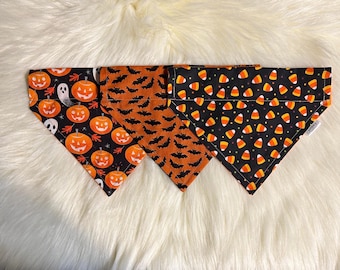 May include: Three Halloween-themed dog bandanas. One features orange and black pumpkins with a ghost, another has orange and black bats, and the last one has black and orange candy corn.