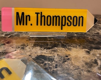 May include: Yellow pencil-shaped desk sign with a pink eraser and black text that reads "Mr. Thompson".
