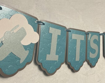 May include: A blue and gray banner with the text "IT'S A" in white letters. The banner is decorated with a white cloud and a silver airplane.