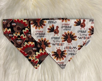 May include: Two Thanksgiving-themed dog bandanas. One is black with a brown turkey print and the other is beige with a brown turkey print and the text "Gobble 'til you Wobble!"