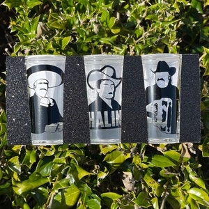May include: Three clear shot glasses with black glitter on the outside. Each glass features a different black and white silhouette of a man wearing a cowboy hat. The middle glass has the word "HALIM" printed on it.