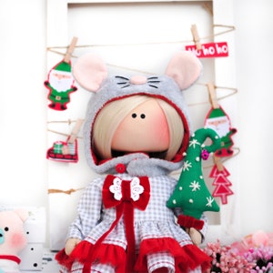 May include: A handmade doll dressed in a gray and white plaid dress with a red bow and a gray hooded coat with mouse ears. The doll is holding a green felt Christmas tree with white snowflakes.