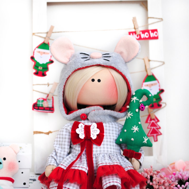 May include: A handmade doll dressed in a gray and white plaid dress with a red bow and a gray hooded coat with mouse ears. The doll is holding a green felt Christmas tree with white snowflakes.