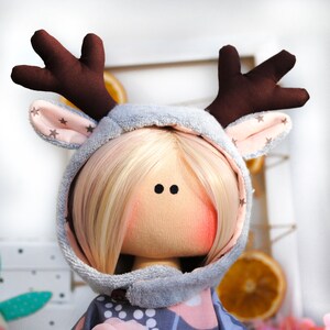 May include: A handmade doll wearing a gray hooded reindeer costume with brown antlers and pink ears. The doll has blonde hair and a light pink face with black eyes.