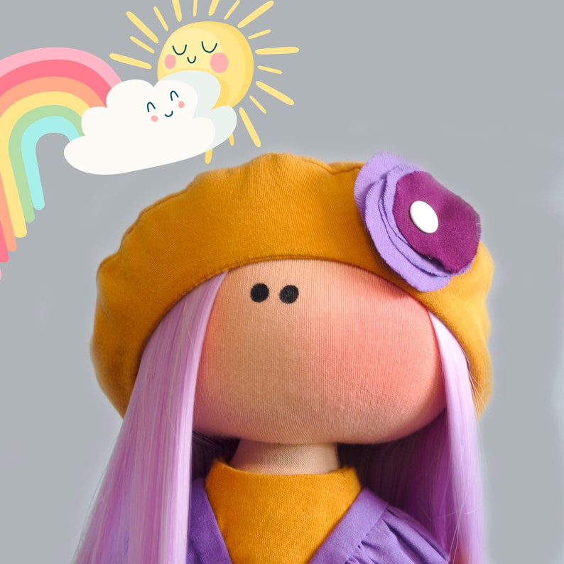 May include: A handmade doll with purple hair, a yellow beret with a purple flower, and a yellow dress. The doll is smiling and has a happy expression. The background is a light gray with a rainbow and a sun.