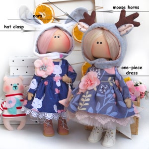 May include: Two handmade dolls wearing dresses and hats. The doll on the left is wearing a blue dress with white bunny print and a white bunny hat with pink ears. The doll on the right is wearing a gray dress with floral print and a gray moose hat with brown antlers. Both dolls have pink flowers on their dresses.