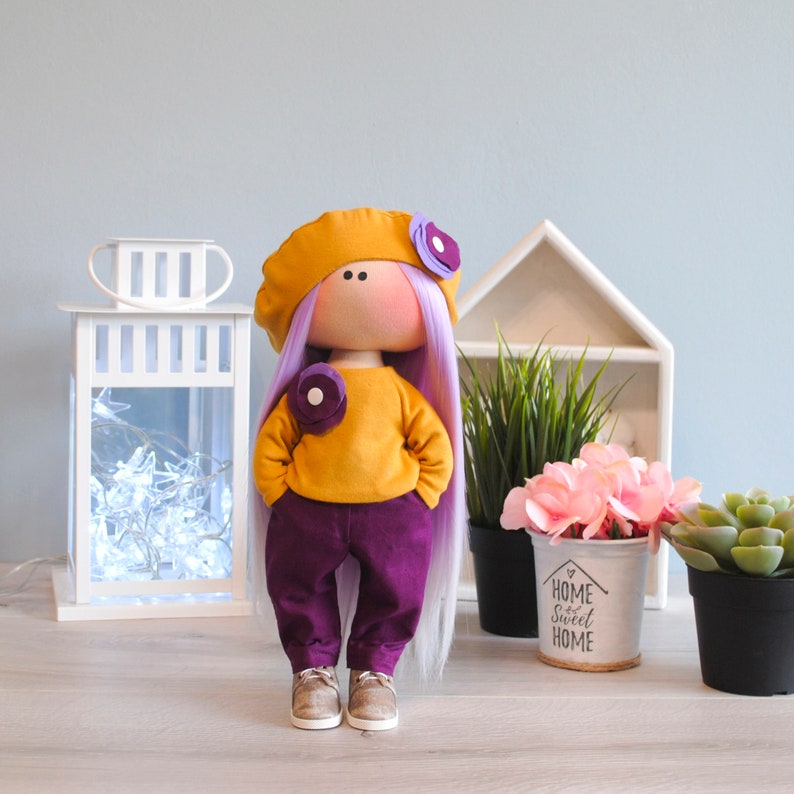 May include: A handmade doll with purple hair, a yellow beret, a yellow sweater, purple pants, and brown shoes. The doll is standing on a wooden surface with a white lantern, a potted plant, and a white planter with the text "Home Sweet Home" on it.