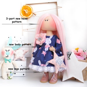May include: A handmade doll with pink hair, a blue dress with a white bunny print, and a pink star. The doll is holding a pink star with silver glitter. The doll is standing on a white surface with a white frame behind it. The image includes the text "3-part new head pattern", "new body pattern", and "new legs pattern".