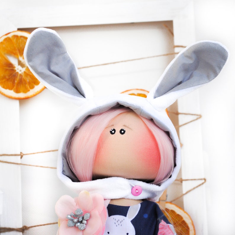 May include: A handmade doll wearing a white bunny costume with gray ears and pink hair. The doll has a pink blush on its cheeks and is wearing a blue and white patterned shirt.