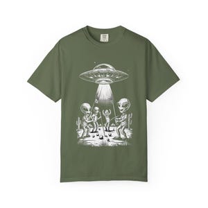 May include: Olive green t-shirt featuring a black and white graphic of aliens playing golf under a UFO. The design includes a detailed illustration of the aliens, golf clubs, golf balls, and desert plants.