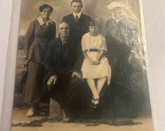 Puede incluir: Una fotografía vintage en blanco y negro de una familia de cinco personas. La familia está posando frente a un cielo nublado. Las dos mujeres llevan vestidos, los dos hombres llevan trajes y la niña lleva un vestido blanco con una cinta negra.