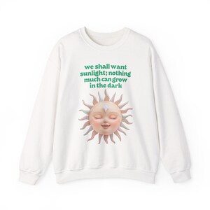 Puede incluir: Sudadera blanca con un gráfico de sol y el texto "we shall want sunlight; nothing much can grow in the dark" en verde. El sol tiene una cara con los ojos cerrados y una estrella en la frente. Los rayos del sol son de color melocotón claro.