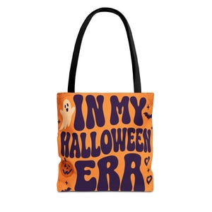 May include: An orange tote bag with black handles featuring the dark purple text IN MY HALLOWEEN ERA in a bold, wavy font. The design includes a small cream-coloured ghost, a carved jack-o'-lantern, a bat, and small heart and star icons.