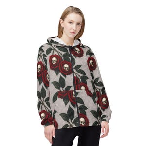 May include: A zip-up hoodie featuring a repeating pattern of red roses with skulls at the center. The roses are set against a light gray background with dark green leaves. The hoodie has a white interior and a black zipper.