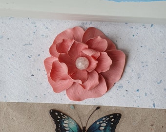 May include: A pink fabric flower with a white crystal center. The flower is on a white background with blue speckles.