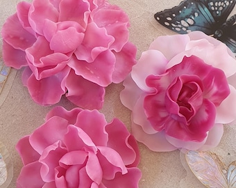 May include: Three pink fabric flowers with ruffled petals. The flowers are arranged on a light brown surface.