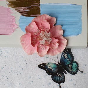 May include: A pink fabric flower with a center of clear and white beads. The flower is on a white surface with a blue and brown painted background. A blue and green butterfly is on the surface below the flower.