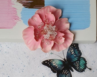 May include: A pink fabric flower with a center of clear and white beads. The flower is on a white surface with a blue and brown painted background. A blue and green butterfly is on the surface below the flower.