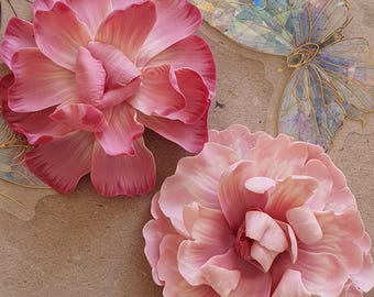May include: Close-up of two large, pink artificial flowers with multiple petals. The flowers are in varying shades of pink, with a textured appearance. Decorative butterfly accents with gold outlines are also visible.