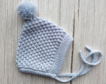 May include: A light blue knitted baby hat with a white pom pom on top and a matching tie string.