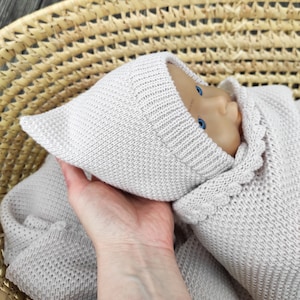 Op de afbeelding: Een lichtbeige gebreide baby muts en bijpassende outfit met een gerimpelde rand. De muts heeft een puntige punt en de outfit heeft lange mouwen. De baby draagt de outfit en de muts.