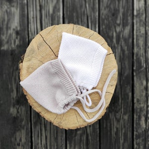 Op de afbeelding: Twee gebreide baby mutsjes, een witte en een lichtbruine, worden getoond op een houten oppervlak. De mutsjes hebben een eenvoudig, klassiek ontwerp en zijn vastgebonden met touwtjes.