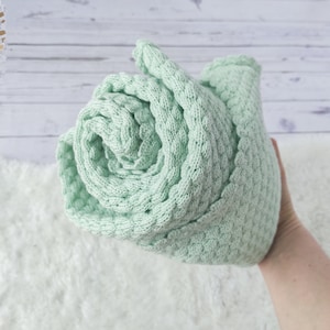 Peut inclure: Une couverture pour bébé en tricot doux de couleur vert clair roulée. La couverture a une texture en nid d'abeille.