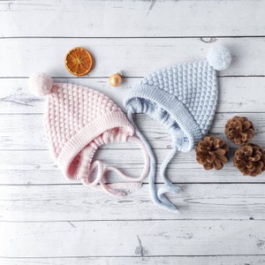 Może przedstawiać: Dwa dziane czapeczki dla niemowląt z pomponami, jedna różowa, a druga niebieska, na białym drewnianym tle. Czapeczki mają fakturowany wzór dzianiny i są związane dopasowanymi wstążkami.