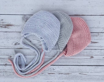 May include: Three knitted baby bonnets in light blue, gray, and pink. Each bonnet has long ties for securing under the chin. The bonnets are displayed on a white wooden surface, showcasing their soft texture and color variations.