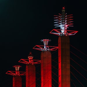 Op de afbeelding: Vier rood verlichte torens tegen een donkere nachthemel. Elke toren heeft een unieke topstructuur met rode en witte lichten. De torens zijn verbonden door rode kabels, wat een opvallende architecturale weergave creëert.