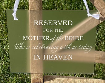 Może przedstawiać: Przezroczysty znak akrylowy z białym tekstem, który brzmi "RESERVED FOR THE MOTHER of the BRIDE Who is celebrating with us today IN HEAVEN". Znak jest zawieszony na białej wstążce na drewnianym krześle.