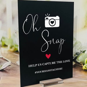 May include: Black sign with white text that says "Oh Snap" with a white camera icon and a red heart. The sign also says "Help us capture the love #YOURHASHTAGHERE".