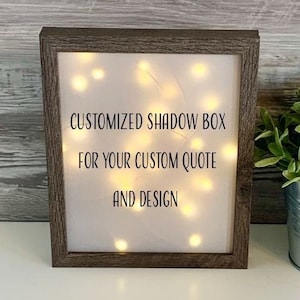 May include: A rectangular, brown-framed shadow box with a white interior illuminated by small, warm-toned lights. The text "CUSTOMIZED SHADOW BOX FOR YOUR CUSTOM QUOTE AND DESIGN" is displayed on the front. A small potted plant is visible on the right.