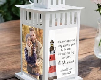 Puede incluir: Linterna de metal blanco con una foto de una pareja y una ilustración de un faro. El texto dice: "Hay quienes traen una luz tan grande al mundo que incluso después de que se hayan ido, la luz permanece. Robert M. Shaw 1934-2024"