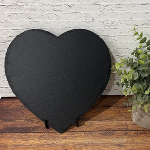 May include: A black heart-shaped slate serving tray with a rustic finish. The tray is on a wooden surface and has two metal stands on the bottom.