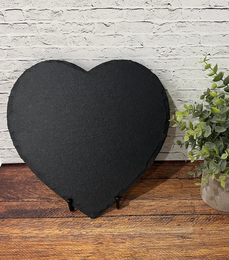 May include: A black heart-shaped slate serving tray with a rustic finish. The tray is on a wooden surface and has two metal stands on the bottom.