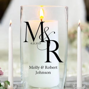 May include: A lit white pillar candle inside a clear glass cylinder, with the initials "M & R" and the date "6.21.2025" printed in black. The names "Molly & Robert Johnson" are printed below. Two white taper candles are on either side.