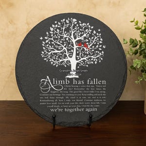 Puede incluir: Placa conmemorativa redonda de pizarra gris oscuro con un diseño de árbol blanco, dos pájaros rojos y texto. El texto incluye un nombre y fechas, junto con un poema. La placa se muestra en un soporte de metal negro.