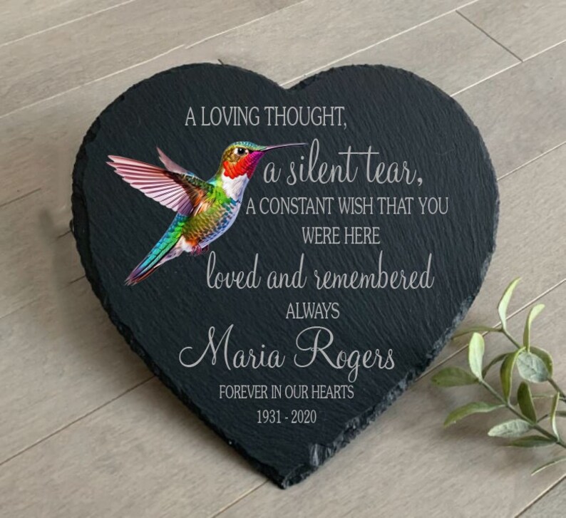 May include: A black heart-shaped slate plaque with a colorful hummingbird illustration. The plaque has the text "A loving thought, a silent tear, A constant wish that you were here loved and remembered always Maria Rogers Forever in our hearts 1931-2020".