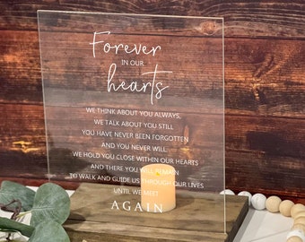 May include: A clear acrylic plaque with the text "Forever in our hearts" and a poem about remembering a loved one. The plaque is sitting on a wooden base with a candle and white beads.