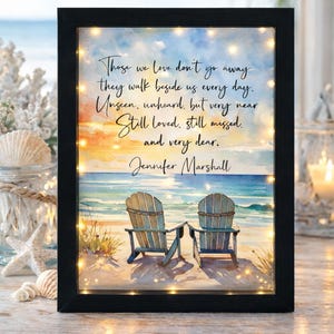 May include: A framed beach scene with two wooden chairs facing the ocean. The artwork includes the text "Those we love don't go away..." and the name "Jennifer Marshall". The frame is black and has small lights around the image.