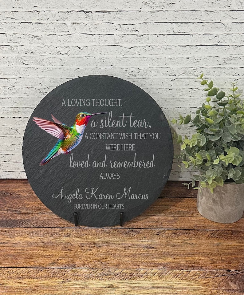 May include: A black slate round plaque with a hummingbird image and the text "A loving thought, a silent tear, A constant wish that you were here loved and remembered always Angela Karen Marcus Forever in our hearts".