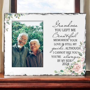 May include: A white slate photo frame with a floral design and a black inscription that reads "Grandma you left me beautiful memories, your love is still my guide. Although I cannot see you you're always by my side 2024 - Ros Tränimer". The frame has a photo of an older couple in the centre.