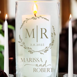May include: A lit white pillar candle inside a clear glass hurricane. The candle is decorated with a floral wreath, initials "MR", the date "3.8.2025", and the names "MARISSA and ROBERT". Two white taper candles flank the hurricane.