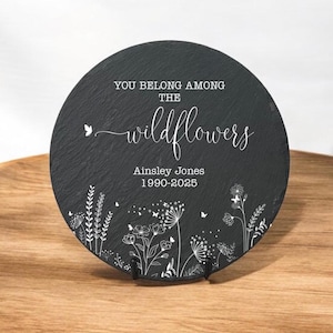 以下が含まれることがあります： 黒いスレートの丸いプレートに、白い文字で「You belong among the wildflowers」と「Ainsley Jones 1990-2025」と書かれています。プレートの縁には白い花の模様が描かれています。